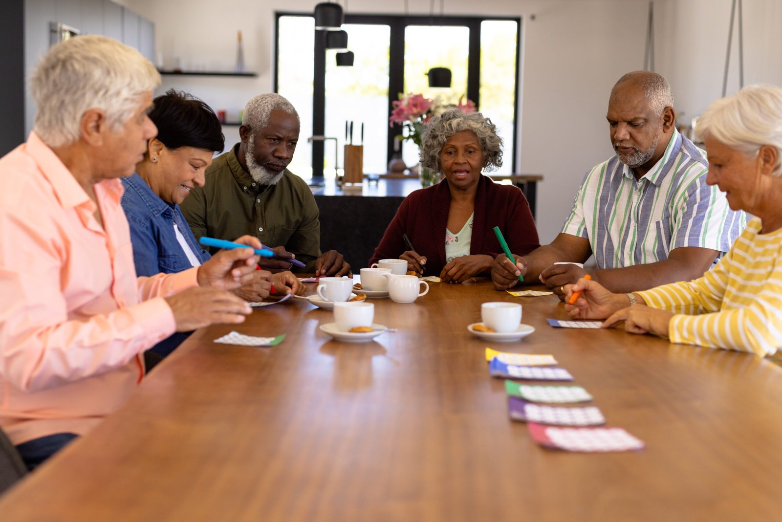 Multiracial senior friends playing bingo while having coffee and cookies at dining table. Nursing home, luck, leisure game, snacks, unaltered, togetherness, support, assisted living, and retirement.